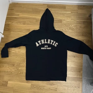 Svart hoodie Athletic North Coast - ( Fick den i present, men den var inte i min storlek).  Svart hoodie med trycket 'ATHLETIC 1979 NORTH COAST' i vitt och beige på bröstet. Klassisk känguruficka framtill och stor huva. Långärmad och i mjukt bomullsmaterial, perfekt för chill dagar. Snygg och enkel stil som funkar till jeans eller joggers. 