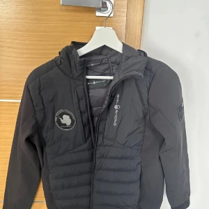 Svart Sail Racing dunjacka med huva - Svart dunjacka från Sail Racing, modell JR Antarctica Hybrid Hood. Jackan har huva, snygga loggor på bröstet och ärmen samt quiltad framsida. Materialmix med nylon, polyester och dun/fjäder. Perfekt för kalla dagar och har en sportig vibe. 2200 ny pris 