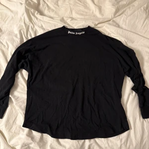 Svart långärmad Palm Angels tröja - Svart långärmad tröja från Palm Angels i 100% bomull. Stor vit logotyp tryckt över ryggen och mindre logga vid halsen. Tröjan har rund hals och ribbade muddar vid ärmslut. Perfekt för dig som gillar streetwear och vill sticka ut med en bold look.