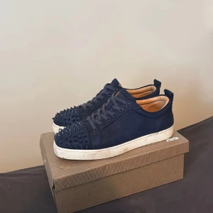 Mörkblå Christian Louboutin sneakers - Snygga mörkblå sneakers från Christian Louboutin med ikoniska röda sulor och coola nitar på tån. Skorna har snörning, rund tå och är tillverkade i mocka med vita platta sulor. Insidan är beige och ger en lyxig känsla. Perfekt för dig som vill sticka ut med exklusiv stil.