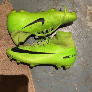 Nike Mercurial gröna fotbollsskor - Säljer ett par grymt snygga Nike Mercurial fotbollsskor i neon-grönt med svart swoosh. Skorna har snörning, platt sula med dobbar och en strumpliknande hög krage för extra stöd. Perfekta för dig som vill sticka ut på planen!