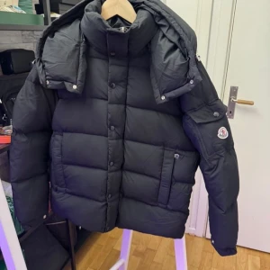 Svart Moncler pufferjacka med huva - Säljer en svart pufferjacka från Moncler med avtagbar huva och hög krage. Jackan har quiltad design, knappstängning och dragkedja, samt Moncler-logga på ärmen. Perfekt för kalla dagar och har praktiska fickor på sidorna. Jackan är helt ny och jag har den i alla storlekar. Mitt pris är 2799 priset är inte hugget i sten 