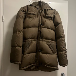 Grön dunjacka från Boomerang XS - Säljer en brun dunjacka från Boomerang i storlek XS. Jackan har huva med snörning, två fickor, samt broderad logga på ärmen. Puffad design och klassisk look, perfekt för kalla dagar.