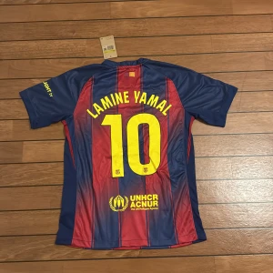 FC Barcelona Lamine Yamal tröja Nike S - Säljer en FC Barcelona matchtröja med Lamine Yamal och nummer 10 på ryggen. Tröjan är kortärmad, tillverkad i Nike Dri-FIT material och har klassiska blå och röda vertikala ränder med gula detaljer. Officiella klubbmärken och sponsorer som Spotify och UNHCR finns tryckta på framsidan.