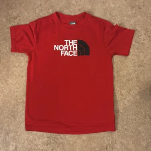 Röd t-shirt från The North Face -  ( xxs/xsSnygg röd t-shirt från The North Face med klassisk logga i vitt och svart på bröstet. T-shirten har rund hals och korta ärmar, perfekt för en avslappnad stil. Materialet känns mjukt och bekvämt mot huden.