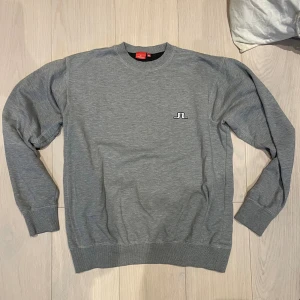 Grå sweatshirt från J.Lindeberg - Snygg grå sweatshirt från J.Lindeberg med broderad logga på bröstet. Tröjan har rund hals, ribbade muddar vid ärmslut och nederkant samt långärmad passform. Perfekt för chill dagar och enkel att matcha med jeans eller joggers.
