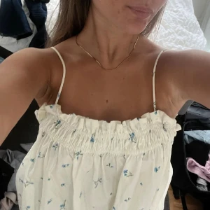 Vit blommig kortklänning med smala axelband - Supersöt vit kortklänning med små blå blommor och smala axelband. Klänningen har en rynkad, elastisk överdel och faller luftigt nedåt. Perfekt för varma dagar och har en lätt, flowy känsla. Passformen är loose och modellen är ärmlös. Helt ny!!