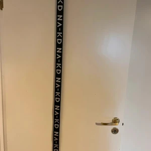 Svart NA-KD bälte med logga - Säljer ett svart bälte från NA-KD med vit logotyp längs hela bandet. Bältet har två silverfärgade ringar som spänne och är gjort i ett slitstarkt syntetmaterial. Perfekt för att ge din outfit en cool streetstyle-vibe.