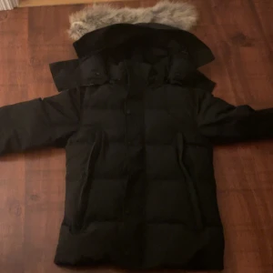 Svart dunjacka från Canada Goose XS - Säljer en svart dunjacka från Canada Goose i storlek XS. Jackan har en fluffig huva med pälsdetalj, långa ärmar och klassisk patch på ärmen. Den är quiltad och har både dragkedja och knappar framtill. Perfekt för kalla vinterdagar.