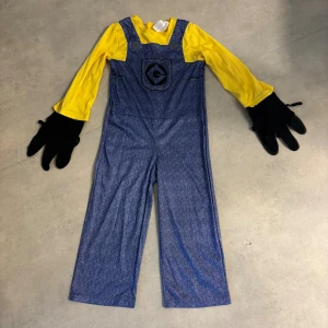 Minion kostym barn strl 104 - Rolig Minion kostym för barn med gula långärmade ärmar, blå hängselbyxor med tryckt logga på bröstet och svarta handskar. Tillverkad i 100% polyester och perfekt för maskerad eller temafest. Den är använd och därför lite sliten på vissa ställen (se bild). Funkar perfekt för barn som vill klä ut sig. 