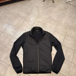 Svart vindjacka med dragkedja - Snygg svart vindjacka från Lindbergh med hel dragkedja framtill och ståkrage. Jackan har en stilren design med slät yta och långa ärmar, perfekt för dig som gillar minimalistisk stil. Passar dig som vill ha en lätt jacka till vardags.