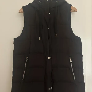 Svart väst med huva och dragkedjor - Svart väst med huva och snörning, perfekt för lager-på-lager. Västen har två sneda fickor med dragkedja och tryckknappar framtill. Quiltad design ger extra värme och stil. 