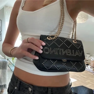 Svart handväska Valentino - Pris kan diskuteras!! Snygg svart handväska från Valentino med quiltat mönster och silverfärgade nitar. Väskan har en kedja i guld som axelrem och stort Valentino-tryck på framsidan. Perfekt storlek för det viktigaste och ger en lyxig vibe till din outfit.