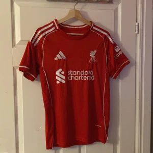 Liverpool Salah #11 röd fotbollströja - Röd Liverpool fotbollströja från Adidas med vita detaljer och tryck. Framsidan har klubbmärke, sponsorlogga och Adidas-logga, baksidan har M. Salah och nummer 11 i vitt. Korta ärmar med vita ränder på axlarna. Materialet är lätt och andas, perfekt för match eller träning.