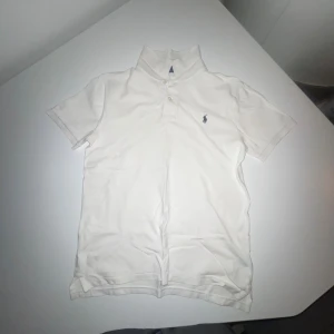 Vit slim fit piké från Polo Ralph Lauren - Vit pikétröja från Polo Ralph Lauren i slim fit-modell. Klassisk design med korta ärmar, krage och två knappar framtill. Ikoniska broderade loggan på bröstet. Tillverkad i mjuk bomull som är skön mot huden. Perfekt för en clean och fräsch look.