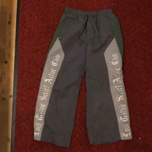 Grå wide byxor Lord Gothic Secret - Grå byxor med bred passform och stora grå paneler längs benen med texten 'Lord Gothic Secret Active Club' i gotisk stil. Byxorna har snörning i midjan och gröna detaljer längs sidorna. Perfekta för dig som gillar streetwear med unik vibe.