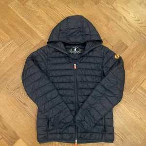 Save The Duck jacka  - Svart pufferjacka med luva från Save The Duck, tillverkad i syntetiskt Plumtech-material som är både andningsbart och vattenavvisande. Jackan har quiltad design, hel dragkedja med orange detaljer. Perfekt för kyliga dagar. Pris kan diskuteras. Skriv om ni har frågor eller om ni vill se mer bilder. 