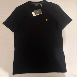 Svart t-shirt från Lyle & Scott - Snygg svart t-shirt från Lyle & Scott i storlek S. Klassisk rund hals och korta ärmar. På bröstet sitter den ikoniska gula fågeln som broderad detalj. T-shirten är tillverkad i mjuk bomull. Tröjan är ny köpt, säljer pga att vi köpte den i fel storlek, tröjan är helt ny, aldrig använd.