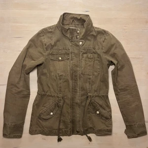 Grön militärjacka från ONLY, strl M - Snygg grön militärjacka från ONLY med hög krage, snörning i midjan och flera fickor med knappstängning. Jackan har dragkedja och tryckknappar framtill, samt långa ärmar. Perfekt för dig som gillar en chill och urban stil. Väldigt fint skick. 