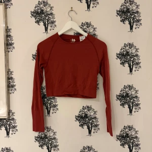 Röd långärmad träningstopp - Säljer en röd långärmad träningstopp från lager 157 med rund hals och ribbade detaljer vid axlarna. Toppen är i ett mjukt och stretchigt syntetmaterial, perfekt för dig som gillar sportig och clean stil. Passformen är tight och den funkar lika bra till jeans som till träningsbyxor.
