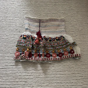 Broderad vit kortkjol med volang - Superfin vit kortkjol med broderade detaljer i flera färger och volangkant. Kjolen har smock i midjan, färgglada broderier, spegelapplikationer och tofsar framtill. Perfekt för dig som gillar boho-stil och unika plagg.