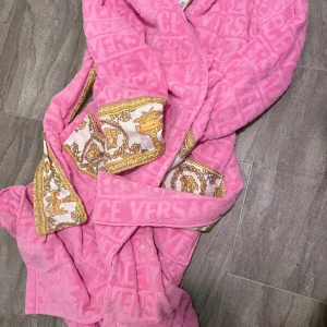 Rosa Versace morgonrock handduk kvalitet med mönster  - Säljer en lyxig rosa morgonrock handduk kvalitet från Versace med ikoniska guldfärgade barockdetaljer och logotypmönster. Jackan har ett bälte i midjan och mjukt, fluffigt material som känns riktigt skönt. Perfekt för dig som vill ha något extra och sticker ut.