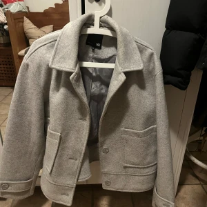 Ljusgrå rock från 157, storlek M - Snygg ljusgrå rock från 157 med klassisk krage och två stora fickor framtill. Jackan har knappar och är helfodrad på insidan. Perfekt för dig som gillar stilrena och enkla plagg med lite boxig passform.