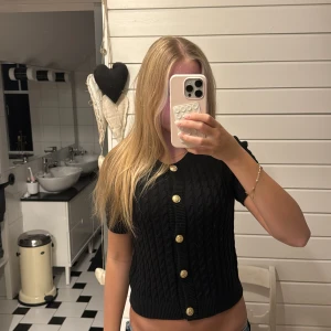 Svart stickad blus med guldknappar - Svart stickad blus med kort ärm och croppad passform. Snygg kabelstickad struktur och dekorativa guldiga knappar framtill. Rund halsringning och ribbade kanter ger en clean look. Perfekt till jeans❣️❣️
