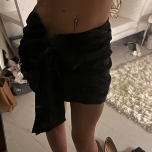 Svart omlottkjol med rosett Gina Tricot - Svart kort omlottkjol från Gina Tricot i storlek 38. Kjolen har en stor rosett på sidan och är tillverkad i ett tunt, lätt material som ger en snygg och trendig look. Perfekt för dig som gillar stilrena plagg med en twist.