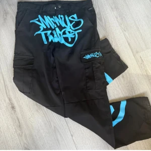 Minus two cargo pants - Svarta cargopants med stora blå graffiti-prints på framsidan, benet och fickan. Byxorna har flera fickor på sidorna och en loose passform. Perfekta för dig som gillar streetwear och vill sticka ut med coola detaljer.