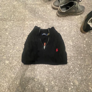 Knitt, Ralph lauren half zipp, stickad tröja - Svart stickad tröja från Polo Ralph Lauren med klassisk kabelstickning och röd broderad logga på bröstet. Tröjan har half zip med läderdetalj på dragkedjan och ribbad krage, ärmslut och nederkant. Perfekt för dig som gillar stilren och tidlös design.