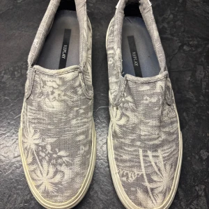 Grå slip-on sneakers från Replay - Gråa slip-on sneakers från Replay med diskret blommigt mönster i vitt. Skorna har en platt sula och är tillverkade i bomullstyg med vita detaljer på hälen där Replay-loggan syns. Perfekta för en avslappnad och stilren look.