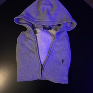 Grå hoodie från Polo Ralph Lauren - Snygg grå hoodie från Polo Ralph Lauren med dragkedja och klassisk huva. Tröjan har lång ärm och den ikoniska Polo-loggan broderad på bröstet. Tillverkad i mjuk bomullsmix som är skön att ha på sig. Perfekt för chill dagar eller när du vill ha en clean look.