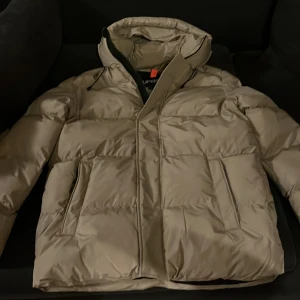 Beige pufferjacka med huva från Superdry - Säljer en beige pufferjacka från Superdry med huva och dragkedja. Jackan har en clean look med stora, quiltade sektioner och två sidofickor. Perfekt för kalla dagar och har en modern, oversized vibe.