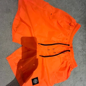 Orange badshorts från Stone Island - Snygga orangea badshorts från Stone Island med svart snörning i midjan och klassisk logopatch på benet. Shortsen har elastisk midja och är tillverkade i lätt polyester med meshfoder. Perfekt för sommaren och strandhäng! Går att skanna och man kommer in på den riktiga hemsidan! 