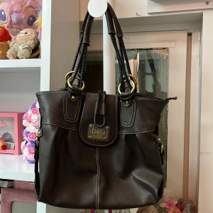 Brun axelväska från D&G i skinn - Snygg brun axelväska från D&G med grova svarta flätade handtag och stora guldfärgade ringar. Väskan är tillverkad i skinn med kontrasterande vita sömmar och har en praktisk dragkedja samt innerfack. Perfekt för dig som gillar klassisk stil eller Stockholm stil med coola detaljer.