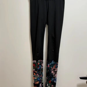 Svarta leggings från Björn Borg med blommigt mönster - Snygga svarta leggings från Björn Borg i storlek 34, med blommigt mönster längst ner på benen. Tillverkade i ett stretchigt syntetmaterial som är perfekt för träning. Modellen är tight och har hög midja för en bekväm passform.