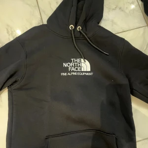 Svart hoodie  - Svart hoodie vit logga och text på bröstet. Klassisk modell med huva, snörning och känguruficka framtill. Perfekt för chill dagar eller när du vill ha en clean streetstyle-look.