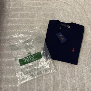 Mörkblå t-shirt från Polo Ralph Lauren - Snygg mörkblå t-shirt från Polo Ralph Lauren med klassisk röd broderad logga på bröstet. T-shirten har rund hals och korta ärmar, tillverkad i mjuk bomull för skön komfort. Perfekt för dig som gillar stilrena och ikoniska plagg.