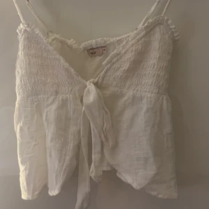 Vit linnetopp med knyt från Nelly - Supersöt vit linnetopp från Nelly i storlek XS. Toppen har smala axelband, smock upptill och en snygg knytning framtill. Materialet är lätt och luftigt, perfekt för varma dagar. Volangdetaljer längs kanten ger en extra gullig touch.
