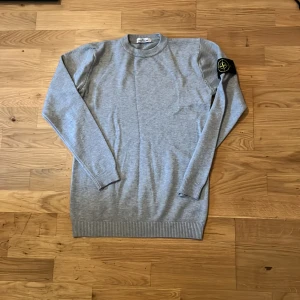 Grå stickad tröja från Stone Island - En helt ny snygg grå stickad tröja från Stone Island med klassisk rund hals och ikonisk logga på vänster ärm. Tröjan har ribbade muddar och är långärmad, perfekt för lager-på-lager under höst och vinter. Pris kan diskuteras vid snabb affär 