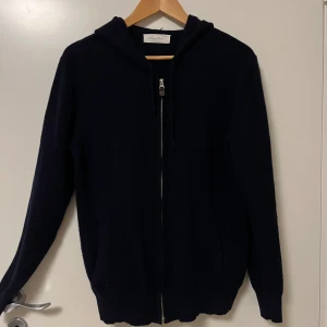 Mörkblå hoodie i kashmir från Sayless - En navy blue hoodie från Sayless i kashmir. Tröjan har dragkedja framtill, huva och långa ärmar. Perfekt för dig som gillar en stilren och enkel look med premiumkänsla.