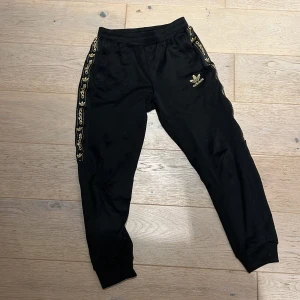 Svarta Adidas mjukisbyxor med guldlogga - Svarta mjukisbyxor från Adidas med guldiga loggor längs sidorna och broderad guldlogga på höften. Byxorna har ribbade muddar vid bensluten och elastisk midja. Materialet är mjukt och skönt, perfekt för chill eller träning.