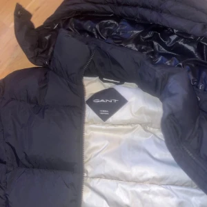 Svart dunjacka från GANT s - Snygg svart dunjacka från GANT i storlek S. Jackan har en quiltad design, huva och dragkedja framtill. Insidan är ljus och jackan är tillverkad av återvunnen nylon och polyester med dunfyllning. Perfekt för kalla dagar och har en clean, modern look. Kan diskutera pris 