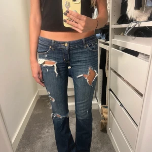 Blå slitna bootcut jeans - Säljer ett par blå jeans med flera slitningar och hål på benen för en edgy look. Modellen är bootcut med låg midja och klassisk femficksdesign. Jeansen har en mörk tvätt och är gjorda i jeansmaterial. Perfekt för dig som gillar streetstyle. Storlek W27, L33. Midjemått 72 cm & innerbenslängd 81 cm