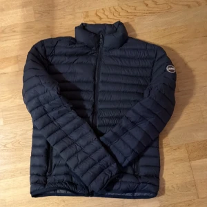 Mörkblå pufferjacka från Colmar - Snygg mörkblå pufferjacka från Colmar med quiltad design och dragkedja framtill. Jackan har långärmade ärmar, ståkrage och två sidofickor med dragkedja. Insidan är grå och har en innerficka med logotyp. Perfekt för kyliga dagar.