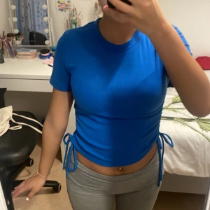 Blå ribbad croppad topp från Zara - Säljer en blå croppad topp från Zara med ribbad struktur och snörning på båda sidorna. T-shirten har rund hals, korta ärmar och är gjord i mjukt syntetmaterial. Perfekt för dig som gillar färg och vill ha en cool detalj med knyt i midjan.