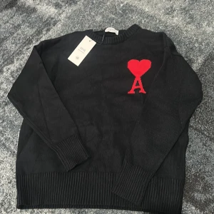 Svart stickad tröja från Ami Paris - Svart stickad tröja från Ami Paris med röd hjärtlogga och bokstaven A på bröstet. Tröjan har rund halsringning, ribbade muddar och är långärmad. Perfekt för dig som gillar stilrena plagg med en twist.
