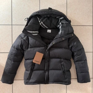 Svart pufferjacka från Burberry - Svart pufferjacka från Burberry med avtagbara ärmar så den kan användas som väst. Jackan har huva med rutigt foder, två dragkedjefickor och knappar framtill. Perfekt för kalla dagar och har en modern, boxig passform.