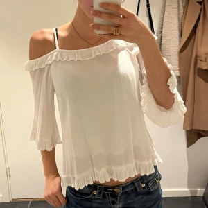 Vit blus med volang och offshoulder - Säljer en söt vit blus med offshoulder och volangdetaljer längs halsringning, ärmslut och nederkant. Blusen har trekvartsärm och är tillverkad i ett lätt, luftigt material som ger en flowy känsla. Perfekt för dig som gillar romantisk och feminin stil.